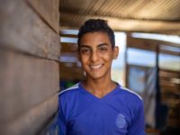 A teen boy in a blue shirt smiles at the camera inside a wooden house.