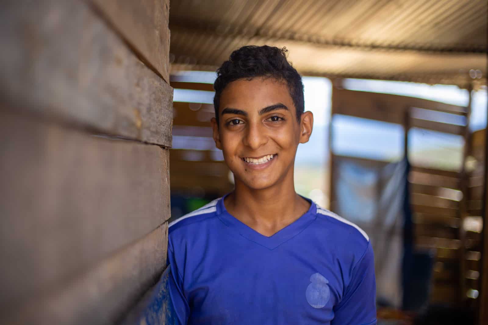 A teen boy in a blue shirt smiles at the camera inside a wooden house.