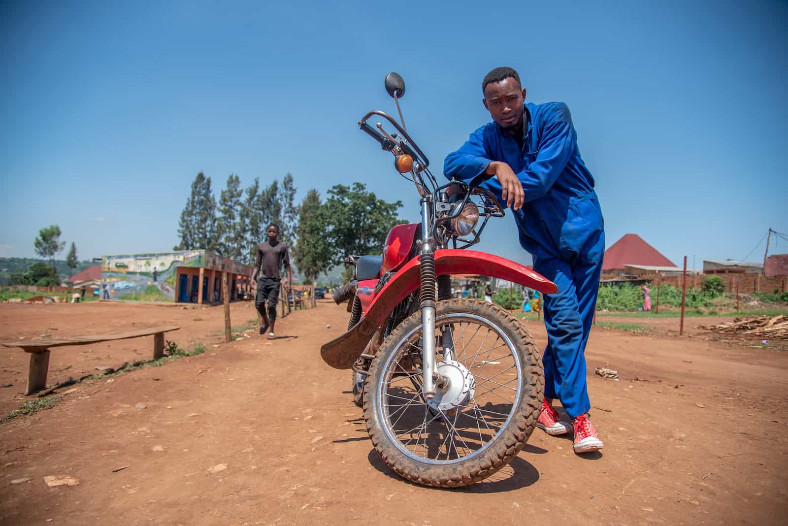 A young man in a blue jumpsuit stands next to a red motorcycle.