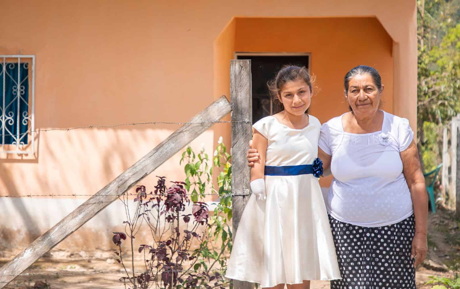 An older woman and a girl with limb difference stand outside an orange house.