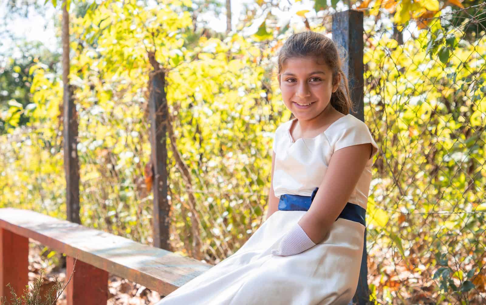 A girl with limb difference sits on a bench outside. 