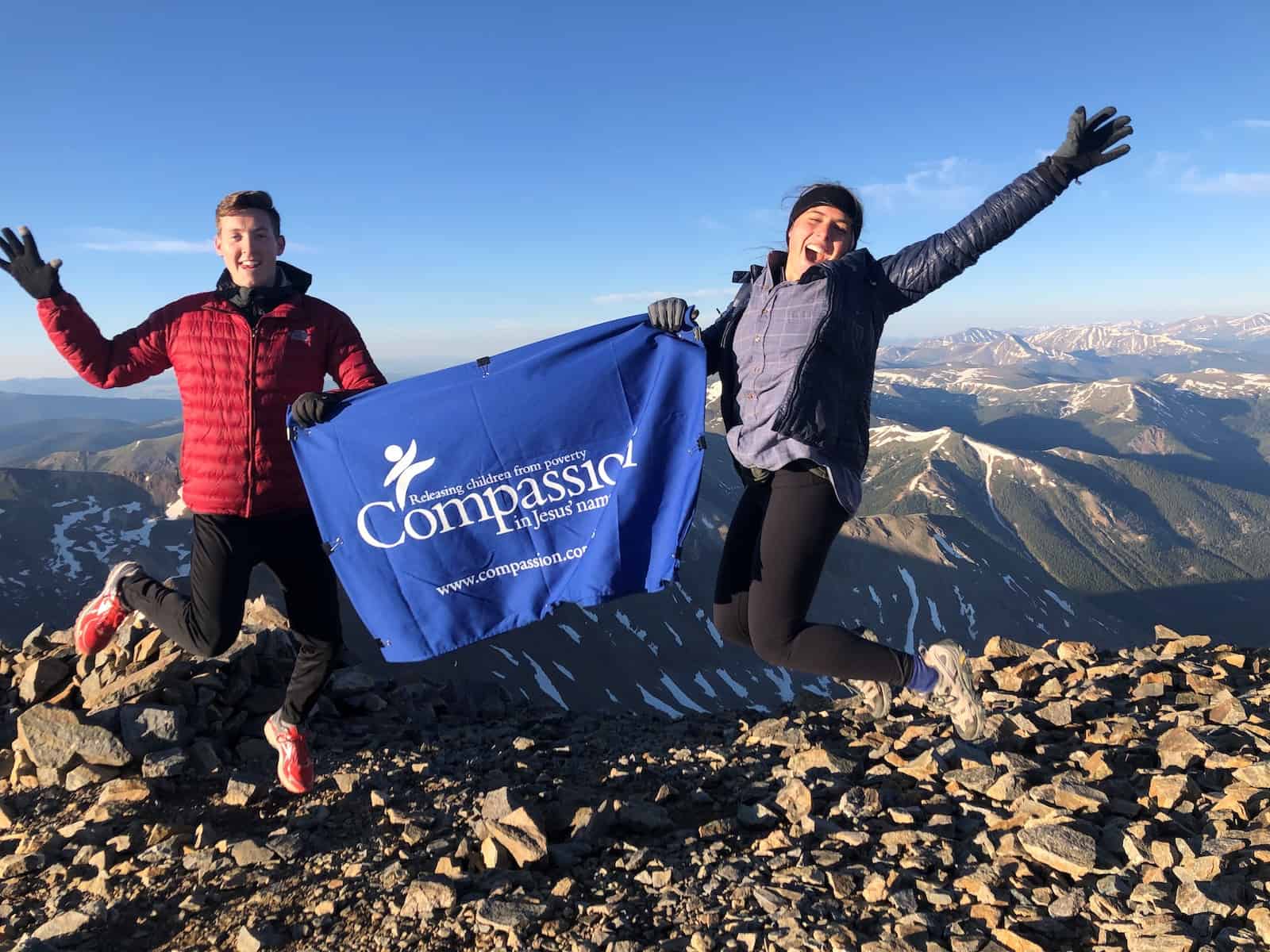 Two people jump in the air, holding a banner that says 'Compassion' on top of a mountain.