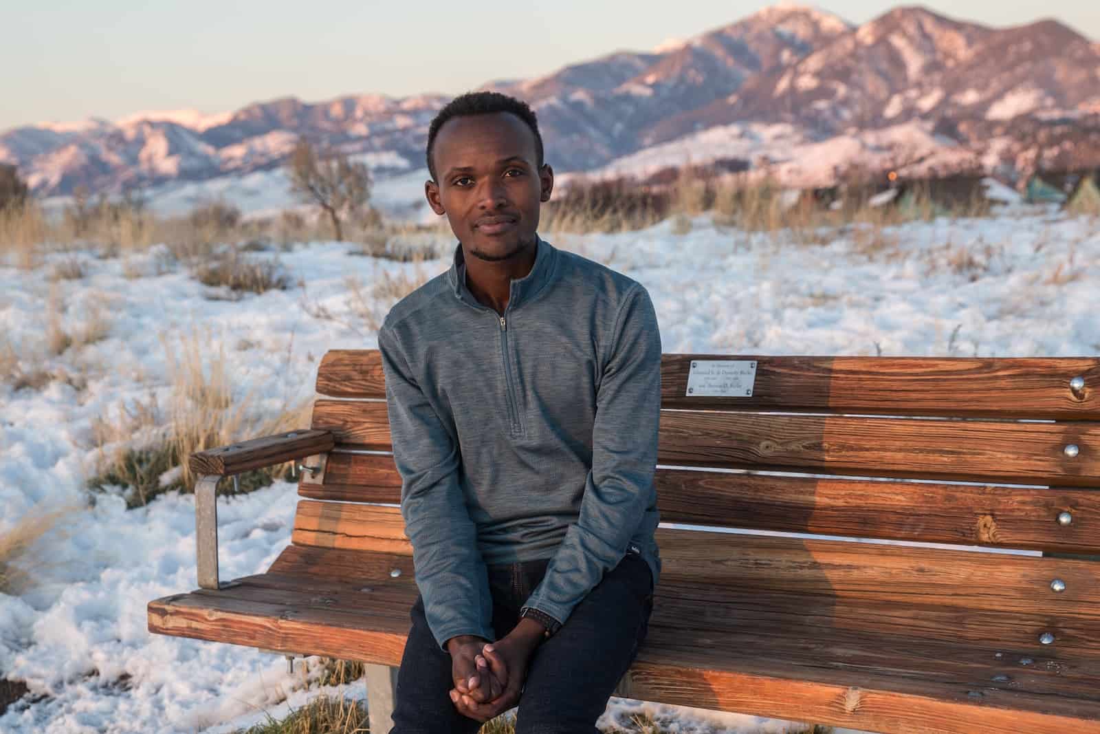A man in a grey sweater sits on a bench in front of snowy hills.