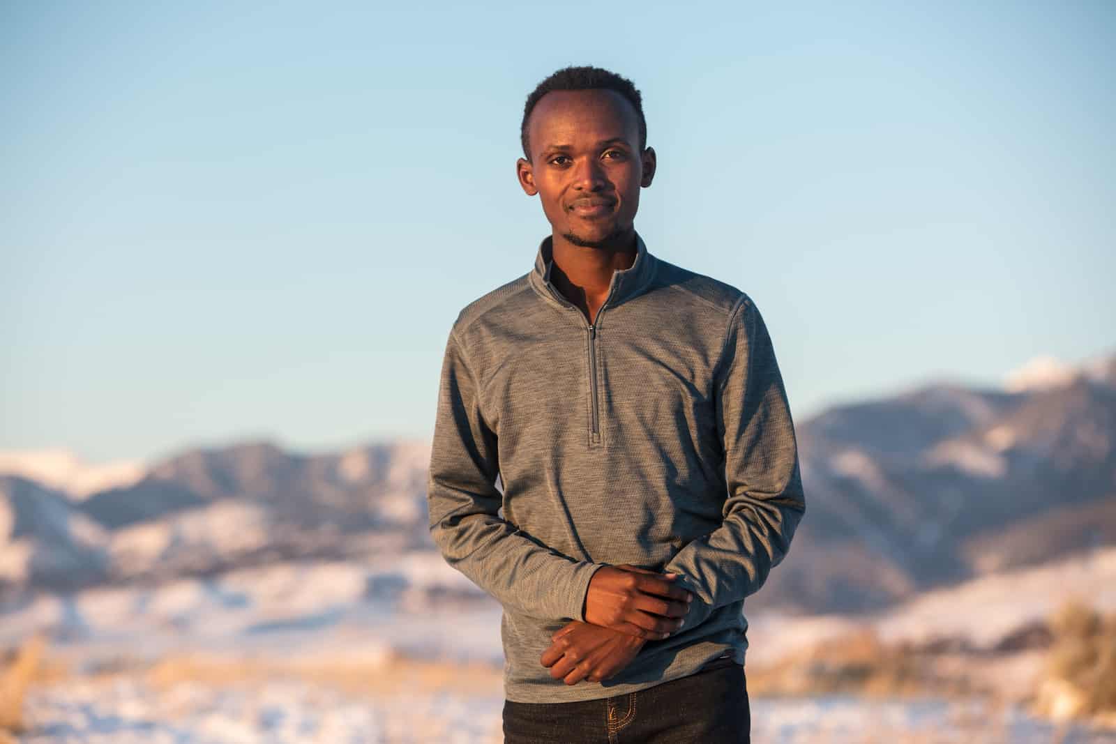 A man stands in a gray shirt in front of snowy hills, smiling at the camera.