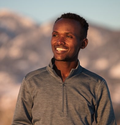 A man in a grey sweater looks off to the side, mountains behind him. 