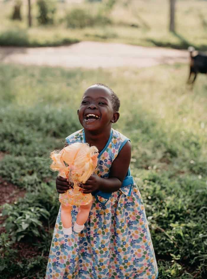 A girl who received a charity gift of heart surgery in a blue floral dress holds a doll and laughs.