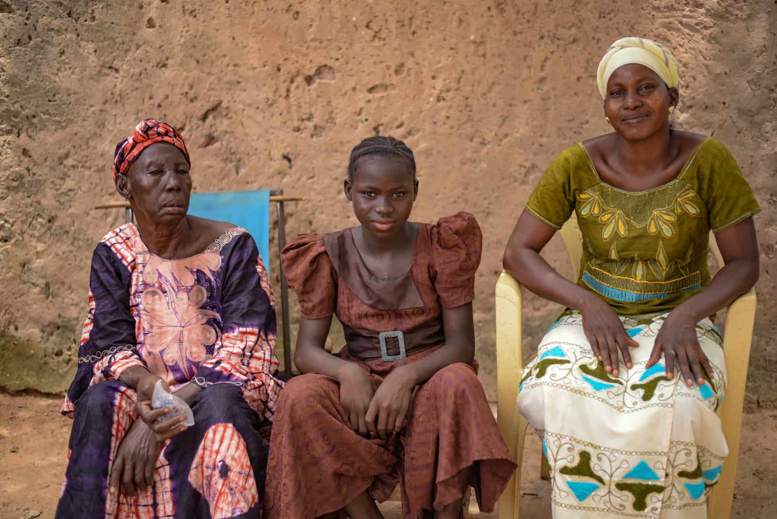 An older women, a girl in a brown dress and a woman in a green and white dress sit in front of a mud wall. 