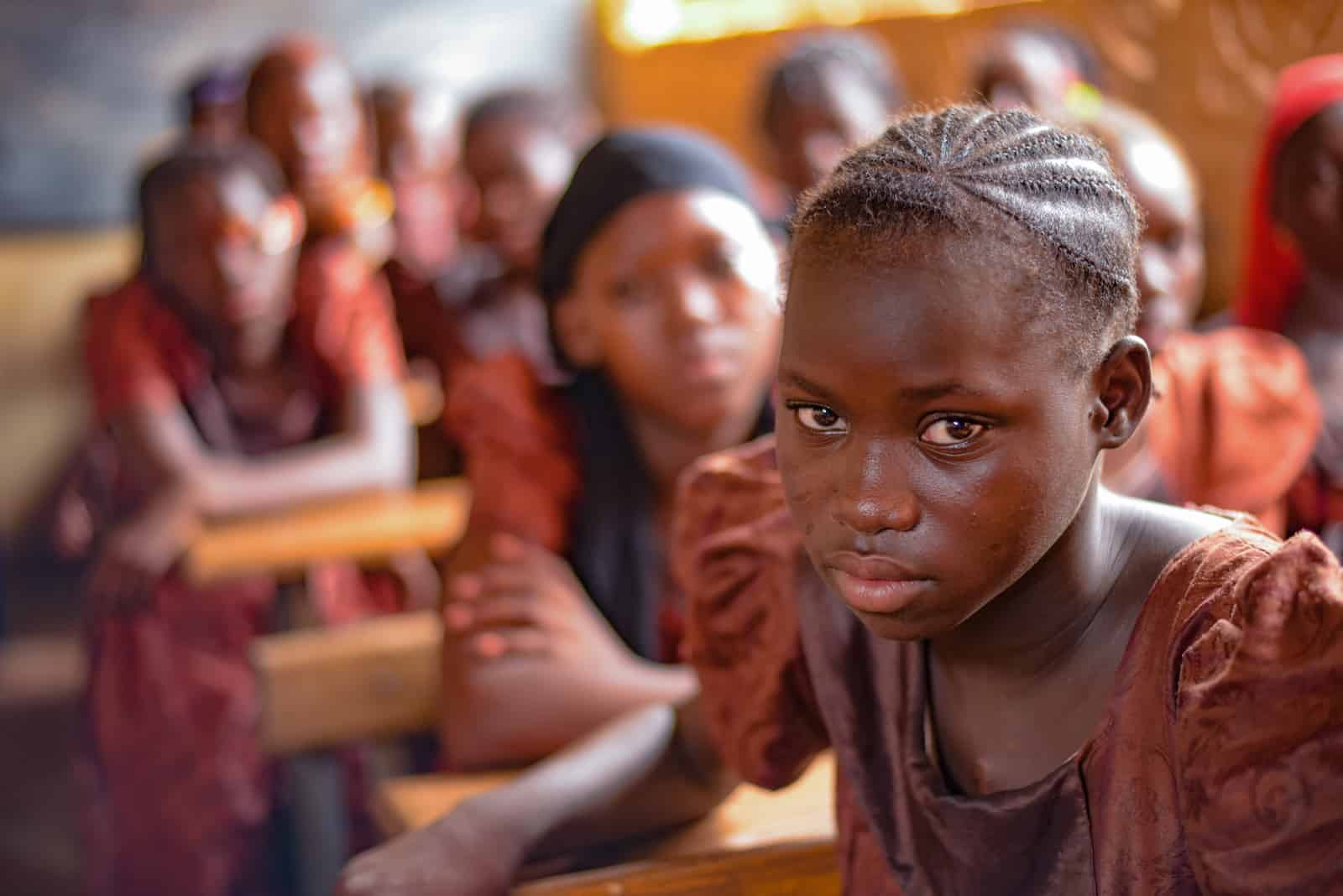 A girl in a brown dress sits in a classroom, looking somber. 