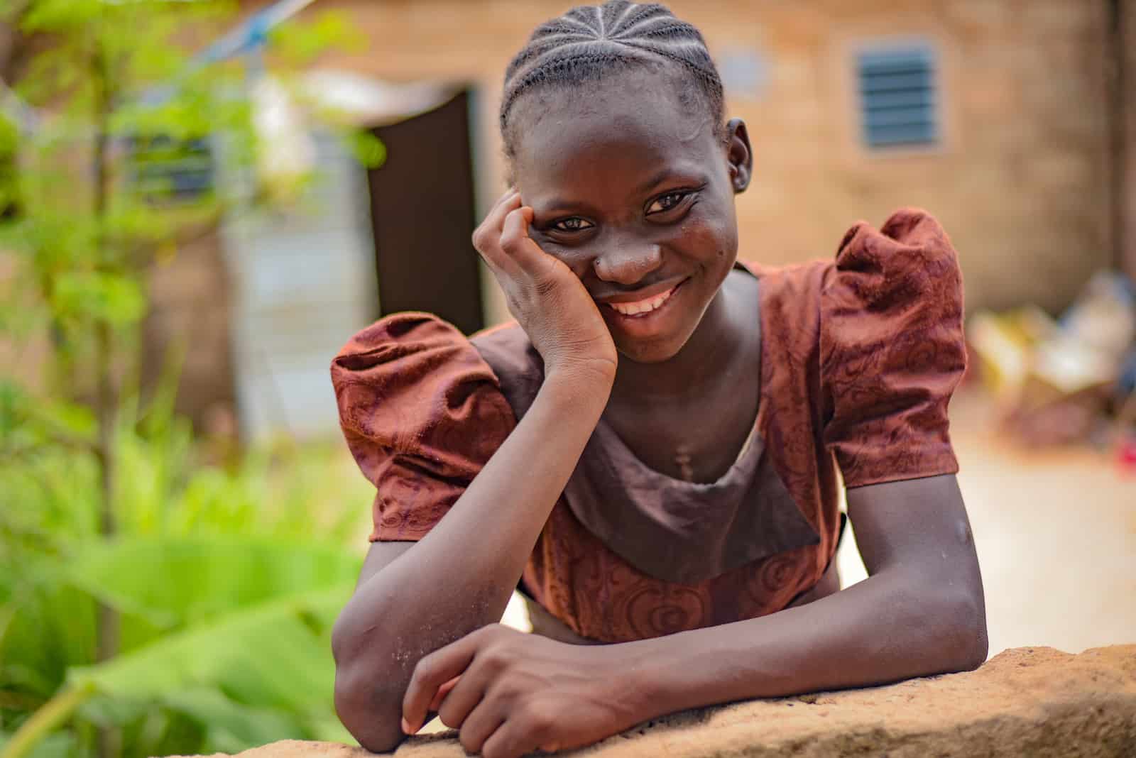 A girl in a brown dress leans on a fence, smiling.