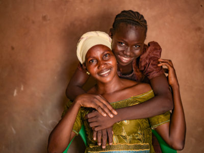 A girl in a brown dress puts her arms around a woman in a green dress and yellow turban, smiling.