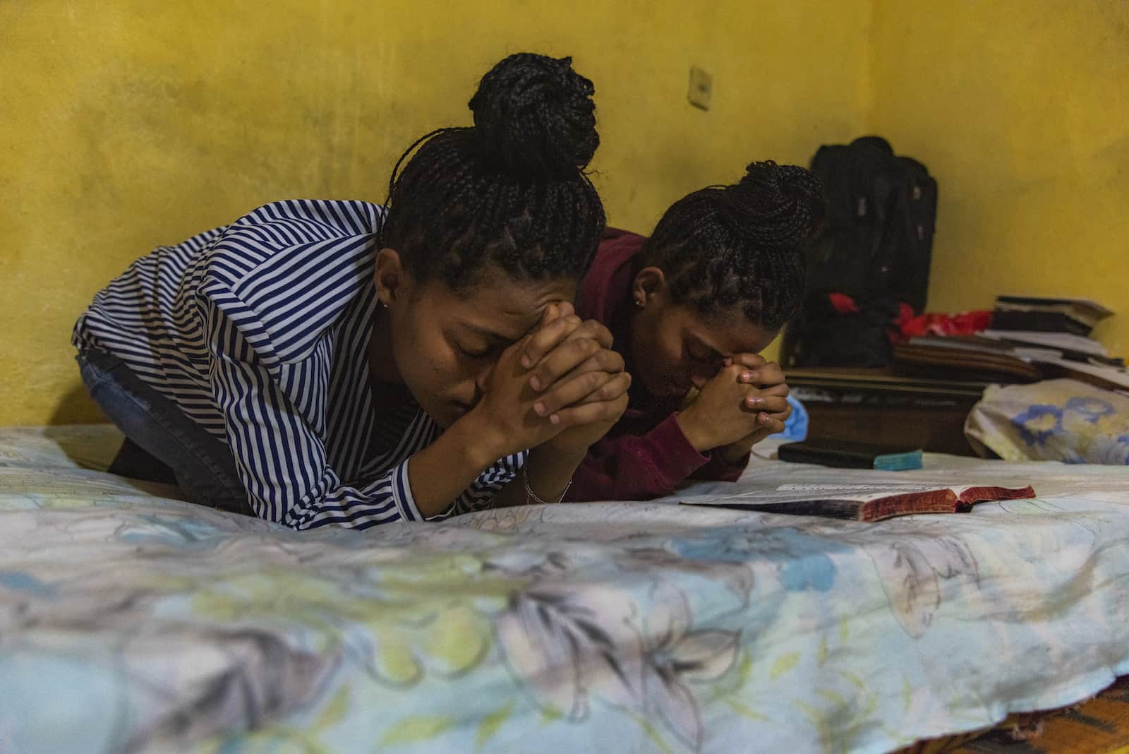 Two girls kneel on a bed, praying.