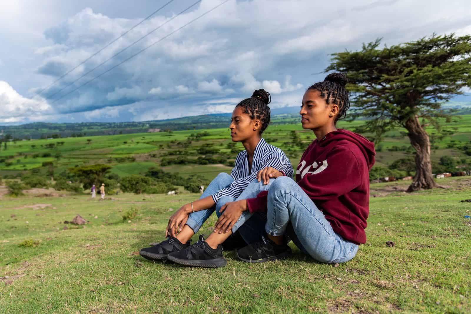 An orphan story: Two teenage girls sit on a hill and look to the distance.