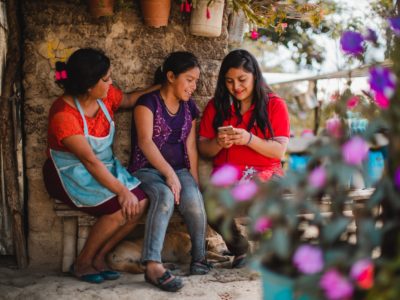 Two women and a girl sit outside on a bench, looking at a cell phone, surrounded by flowers.