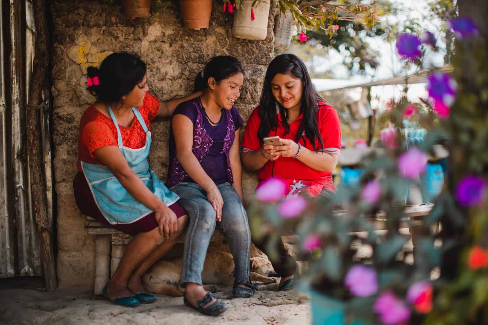 Two women and a girl sit outside on a bench, looking at a cell phone using technology for poverty reduction.