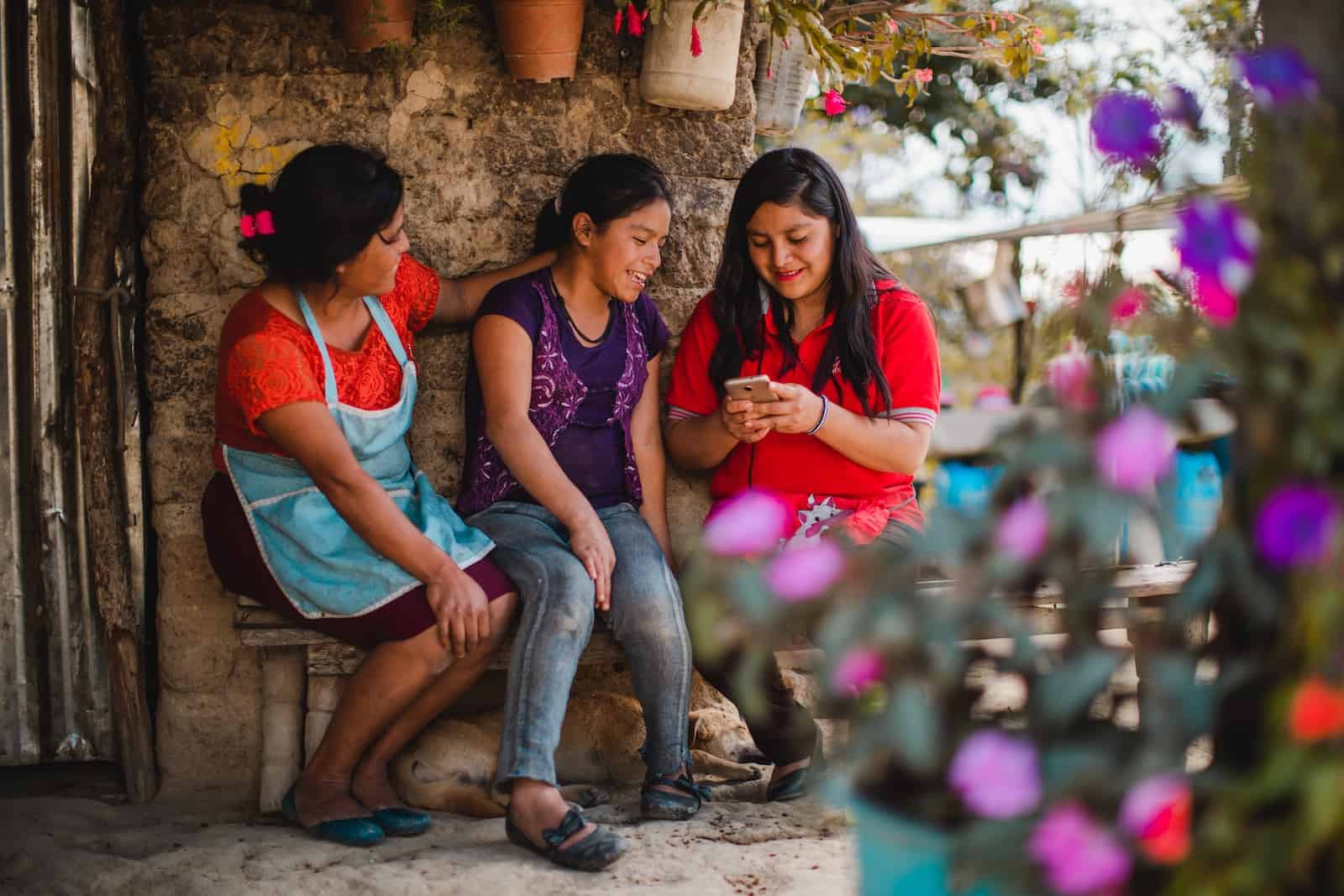 Two women and a girl sit outside on a bench, looking at a cell phone, surrounded by flowers.