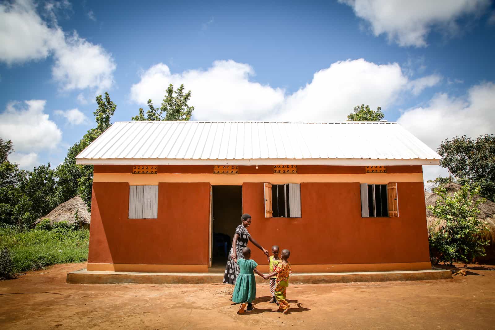 A mother and three children dance in a circle in front of a home with a metal roof. 