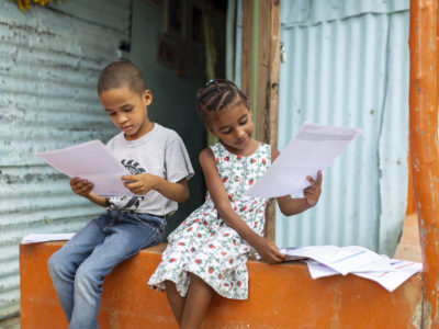 Two children sit outside on a porch, reading letters.