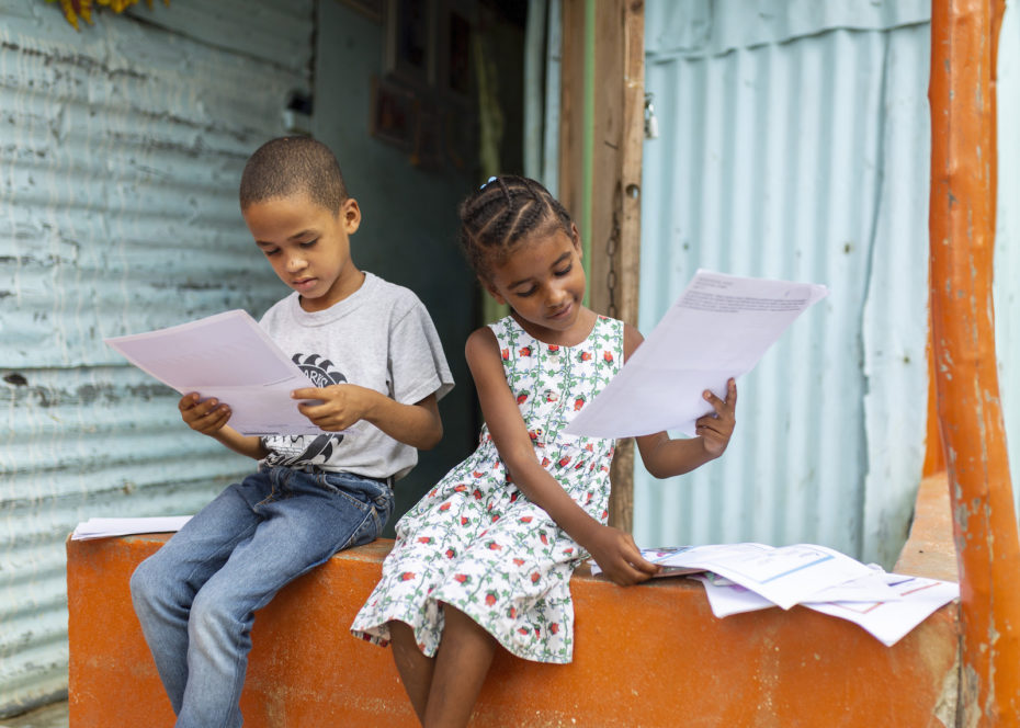 Two children sit outside on a porch, reading letters.