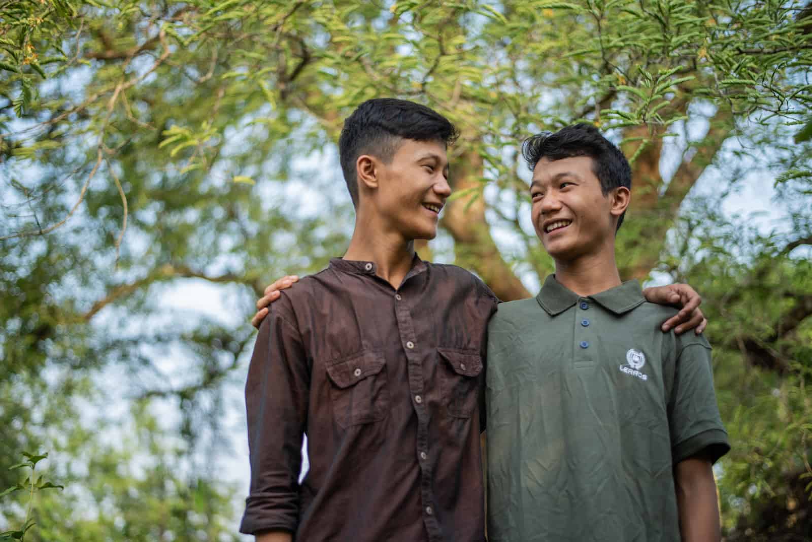 Two teenaged boys smile at each other, with their arms around each other. 