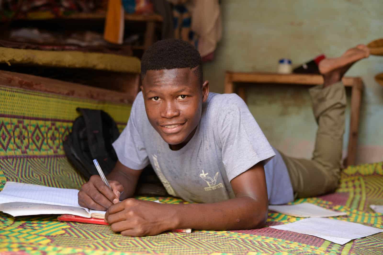 A teenage boy leans over a bed and writes. 