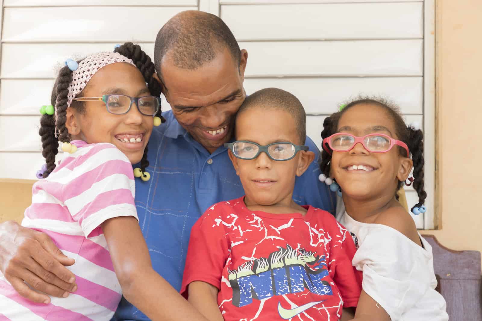 A man sits, hugging three children with glasses. 