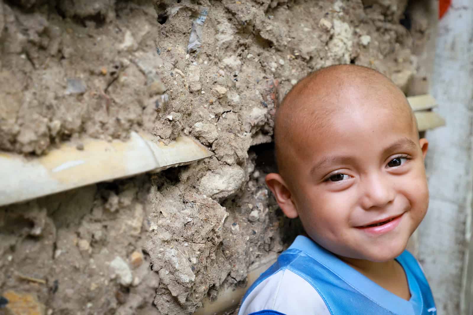 A young boy with no hair leans against a mud wall, smiling.