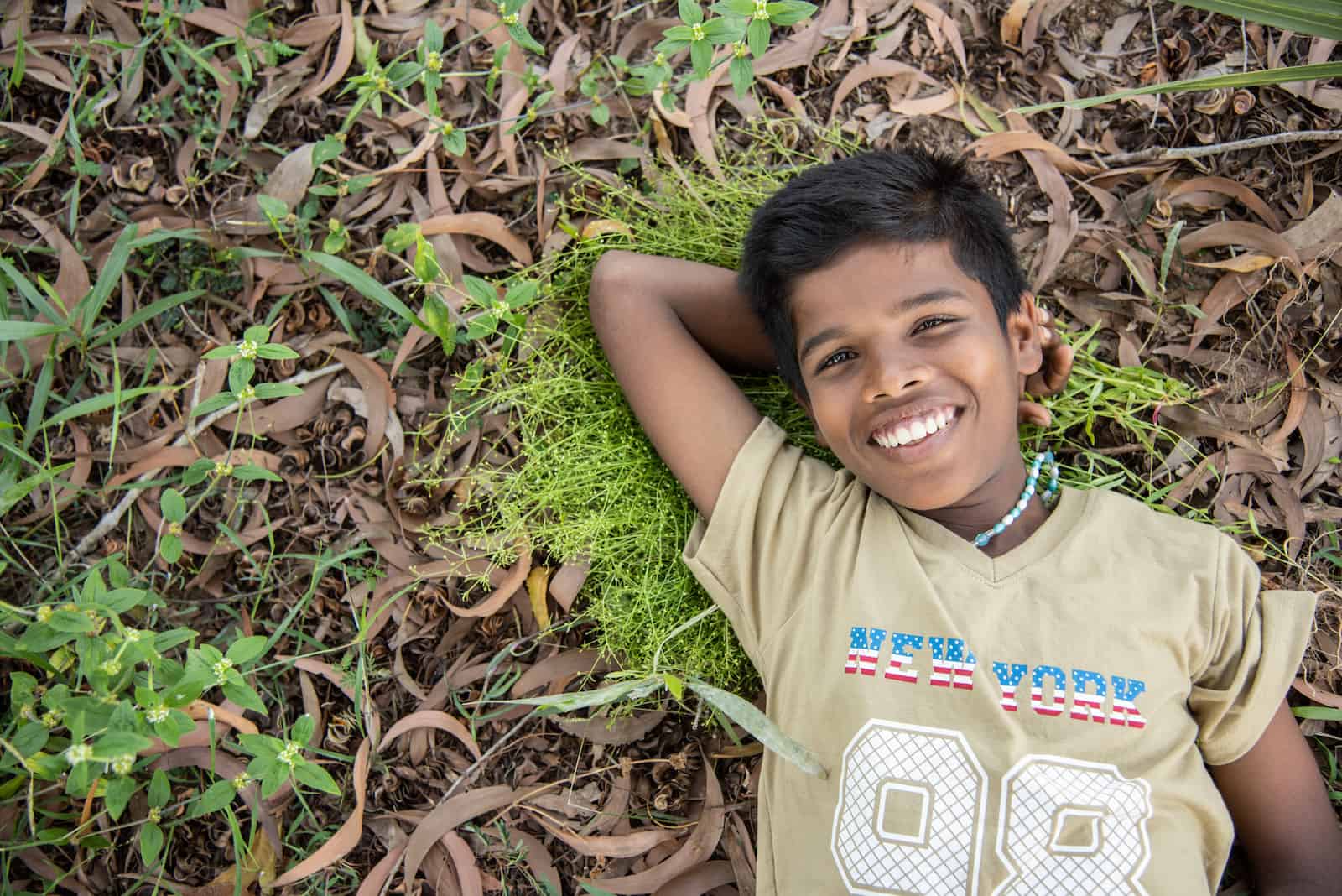 A boy in a tan shirt lays in the grass on the ground, smiling.