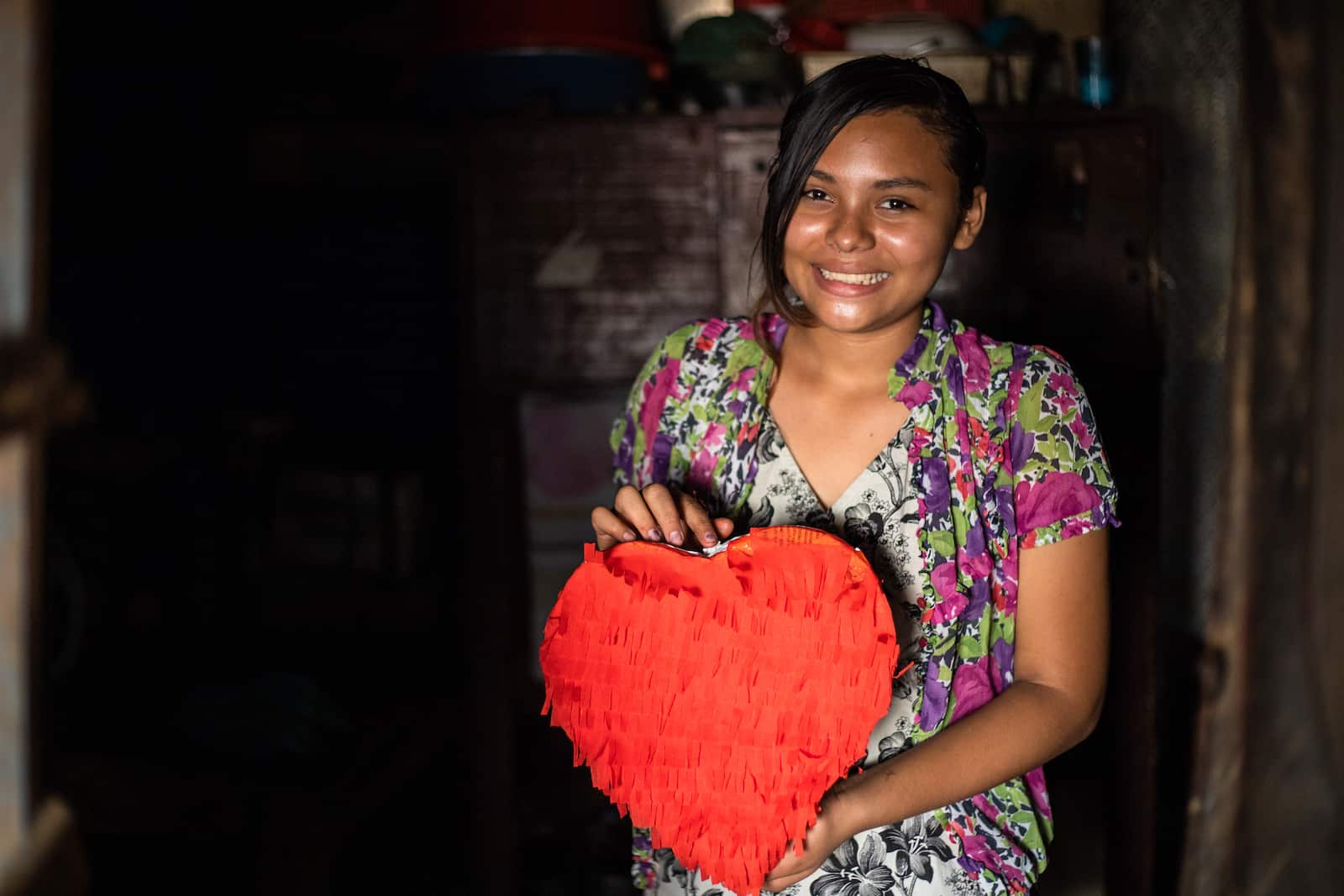 A girl in a floral shirt holds a red heart pinata. 
