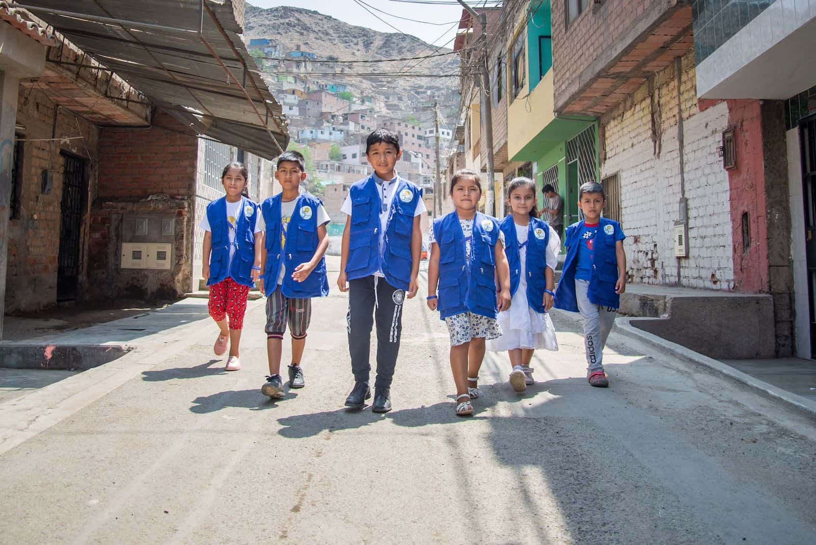 A group of children in blue vests walk down a street in Peru.
