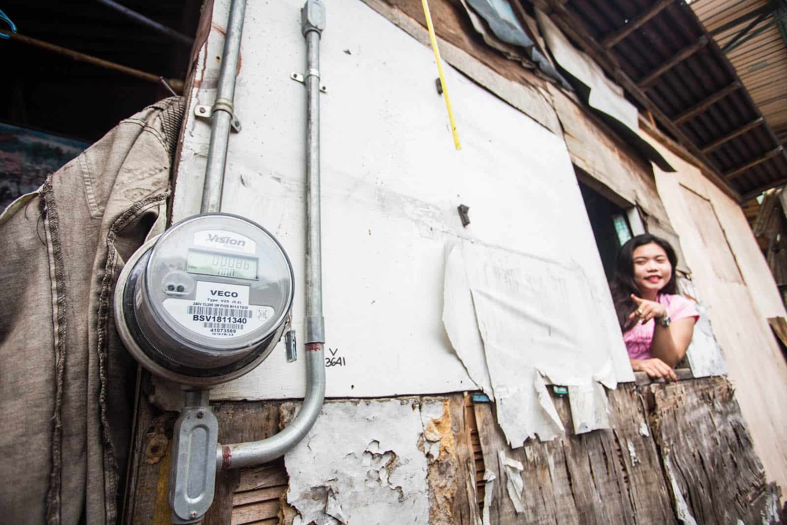 A girls leans out of a window of a makeshift home, pointing at an electrical meter. 