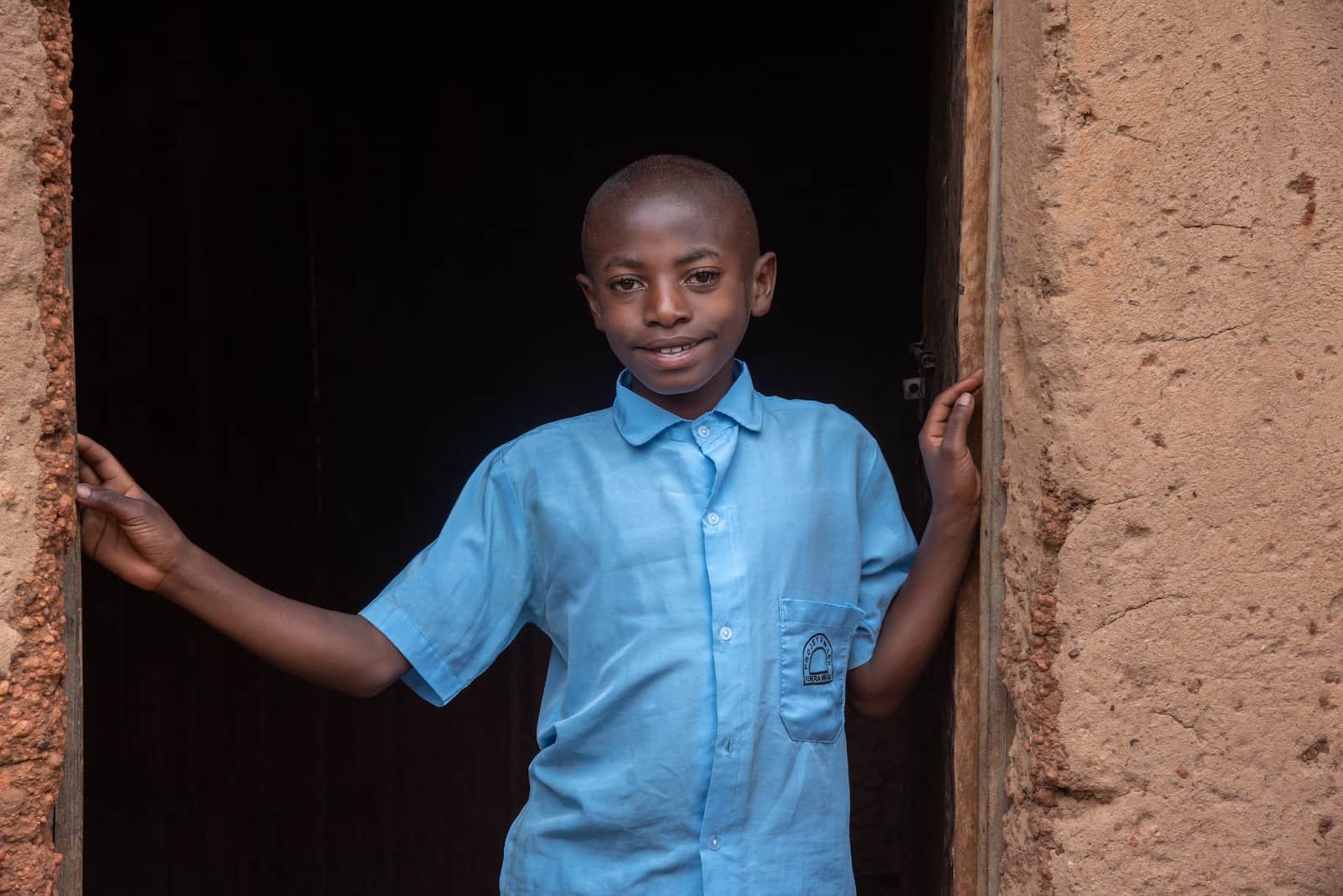 A boy in a blue shirt stands in the door of a mud home.