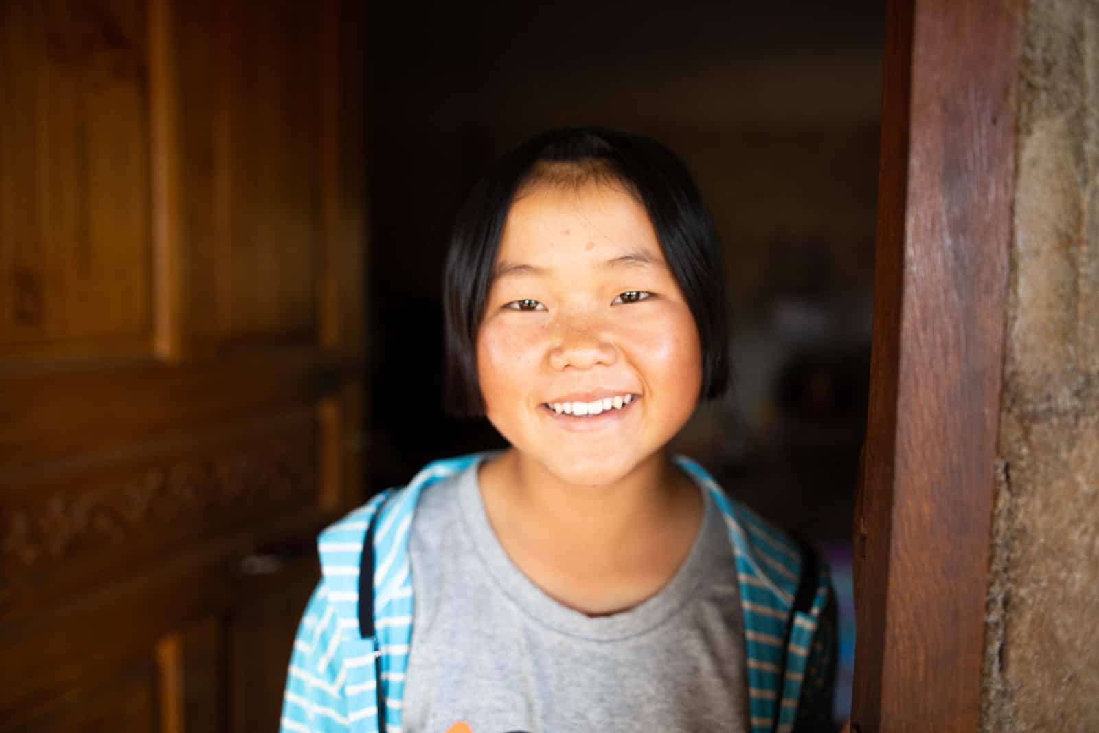 A girl in a grey shirt stands in the doorway of a home, smiling.
