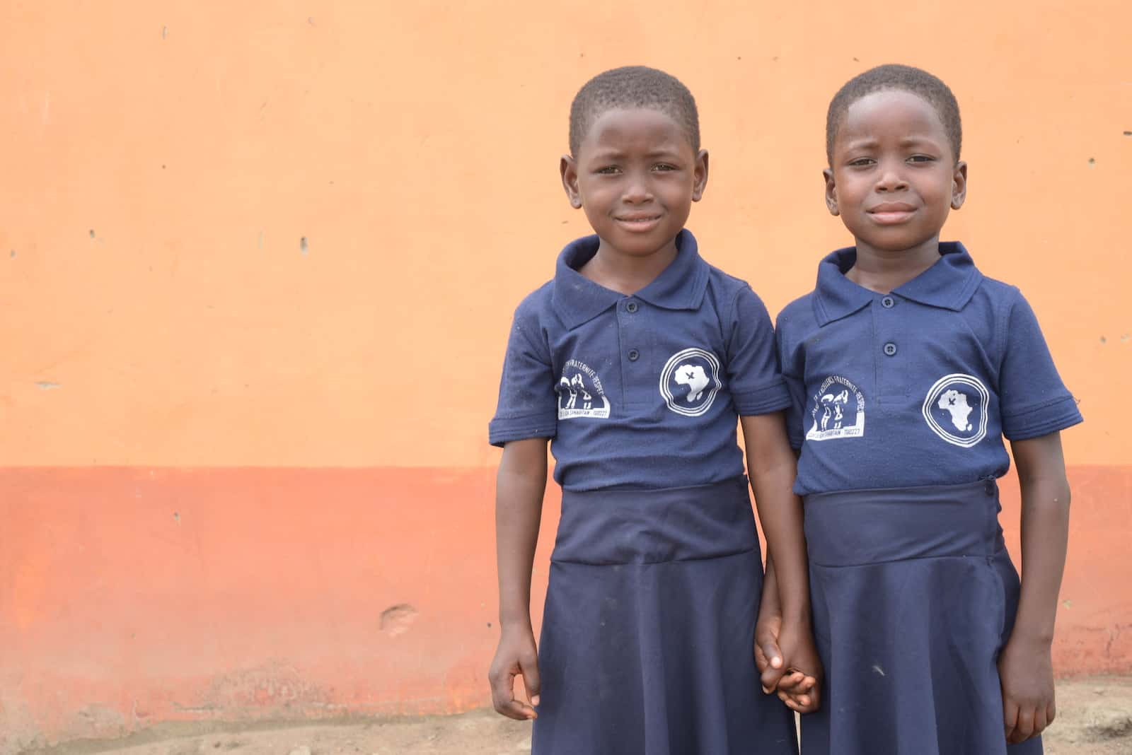 Twin girls in blue school uniforms hold hands, standing in front of an adobe wall. 