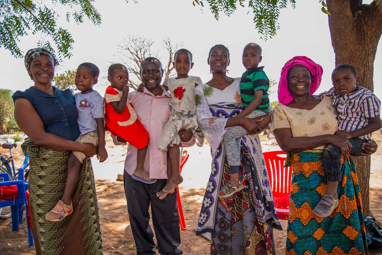 Three women wearing traditional Tanzanian dress and one man hold children in their arms, smiling. 