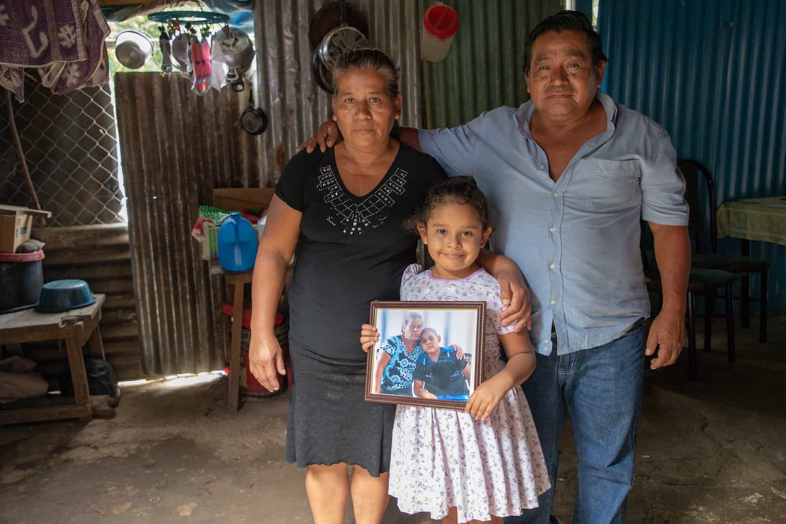 A woman and a man stand behind a little girl holding a picture of a young boy and his grandma. 