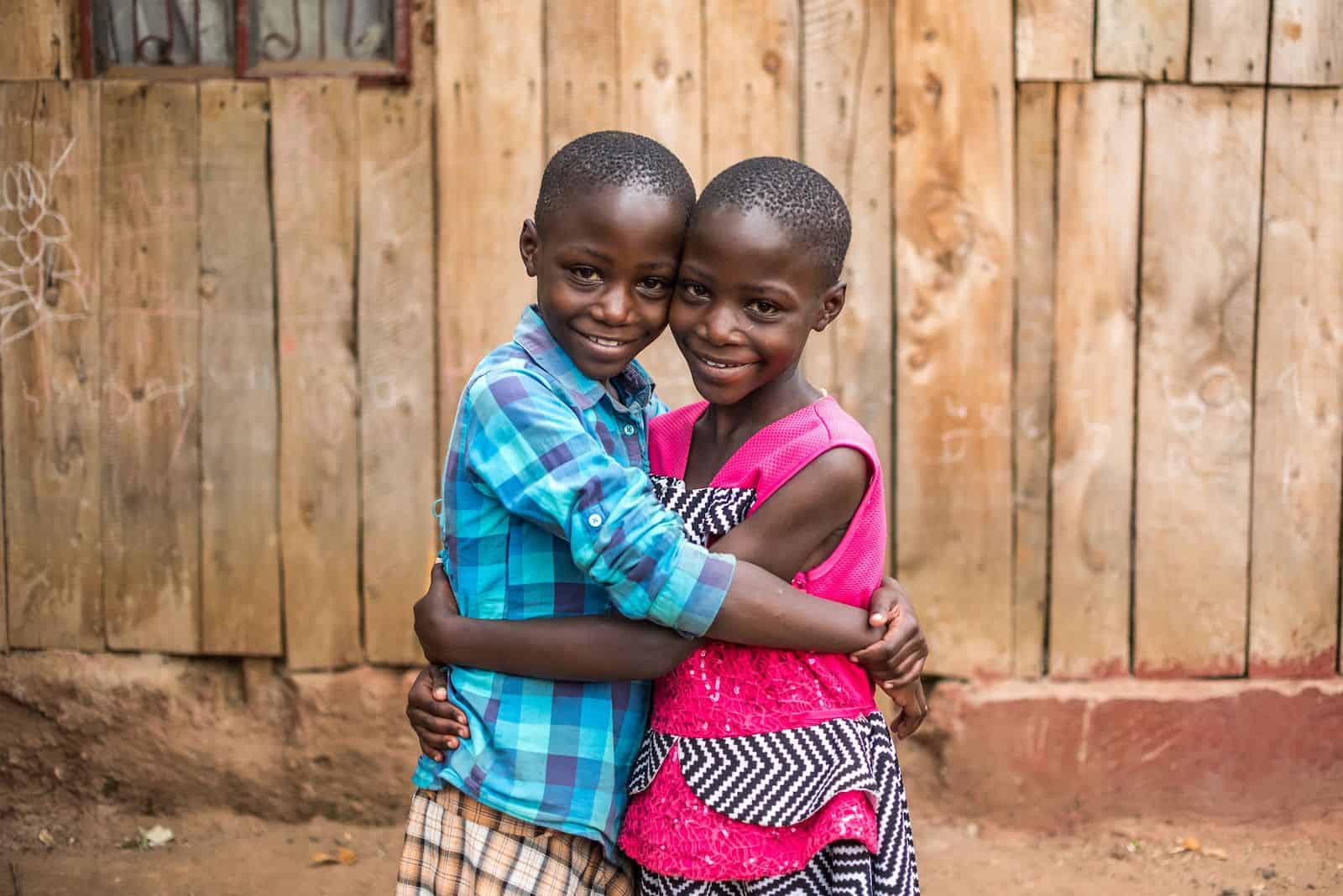 How to Help Kids in Need: Two girls hug each other, standing in front of a wooden home