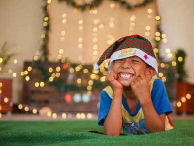 A boy wearing a Santa cap lays on the ground with his face resting in his hands, smiling.