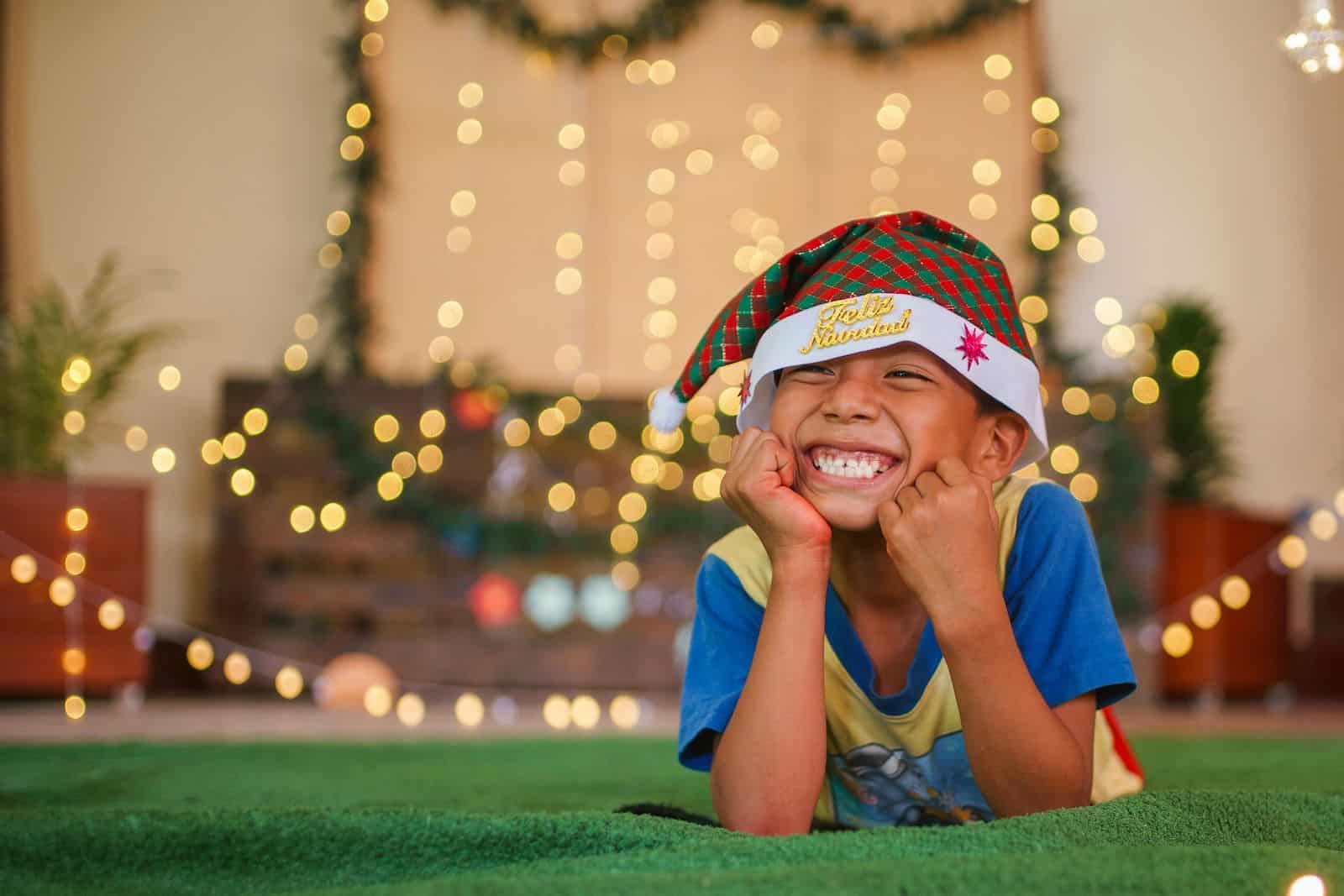 A boy wearing a Santa cap lays on the ground with his face resting in his hands, smiling. 