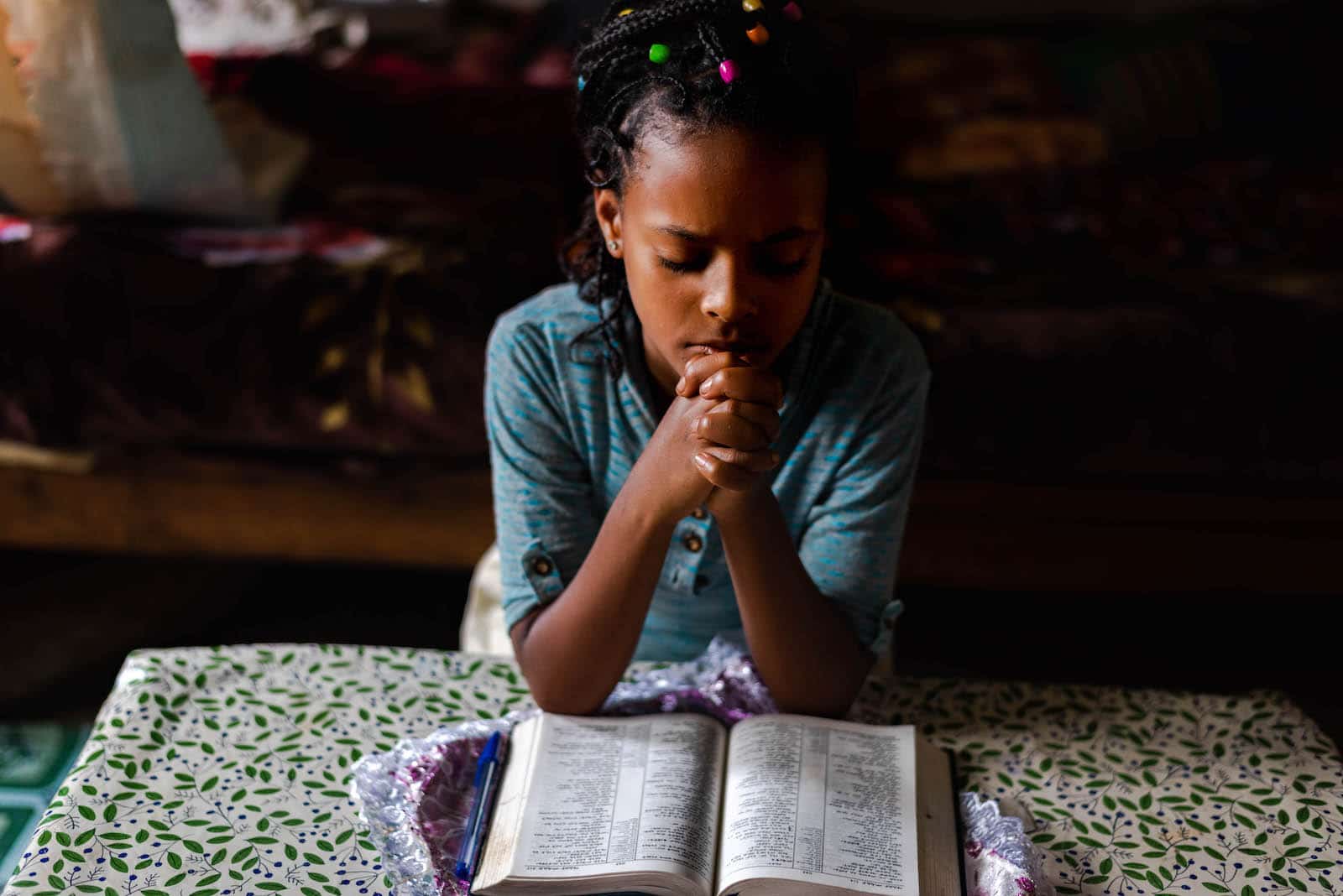 A girl leans on a bed, praying over a Bible.