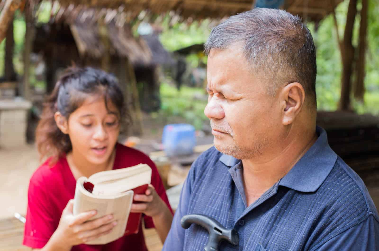 A man listens to a girl read the Bible to him.