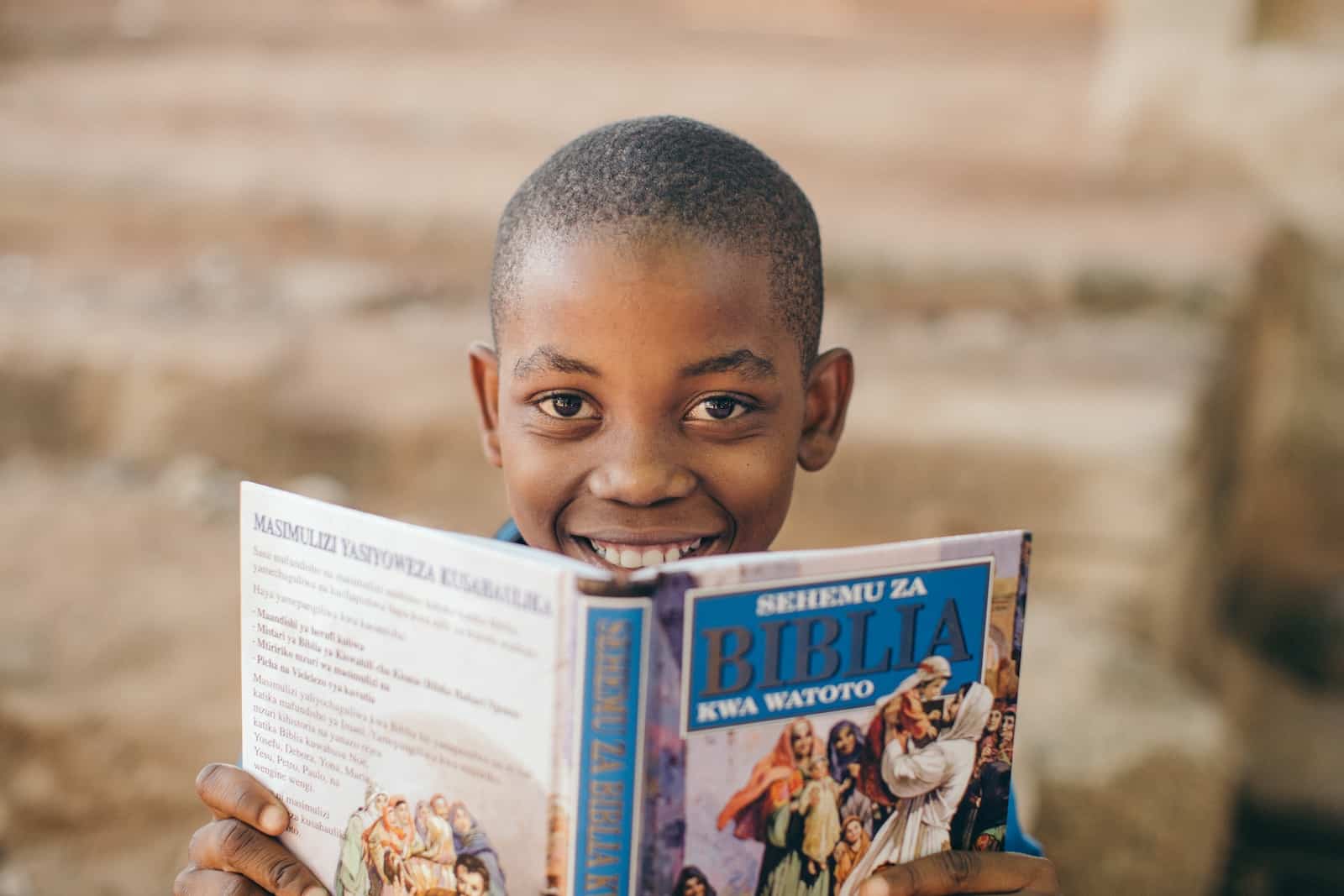 A boy holds an open Bible in front of his face, smiling. 