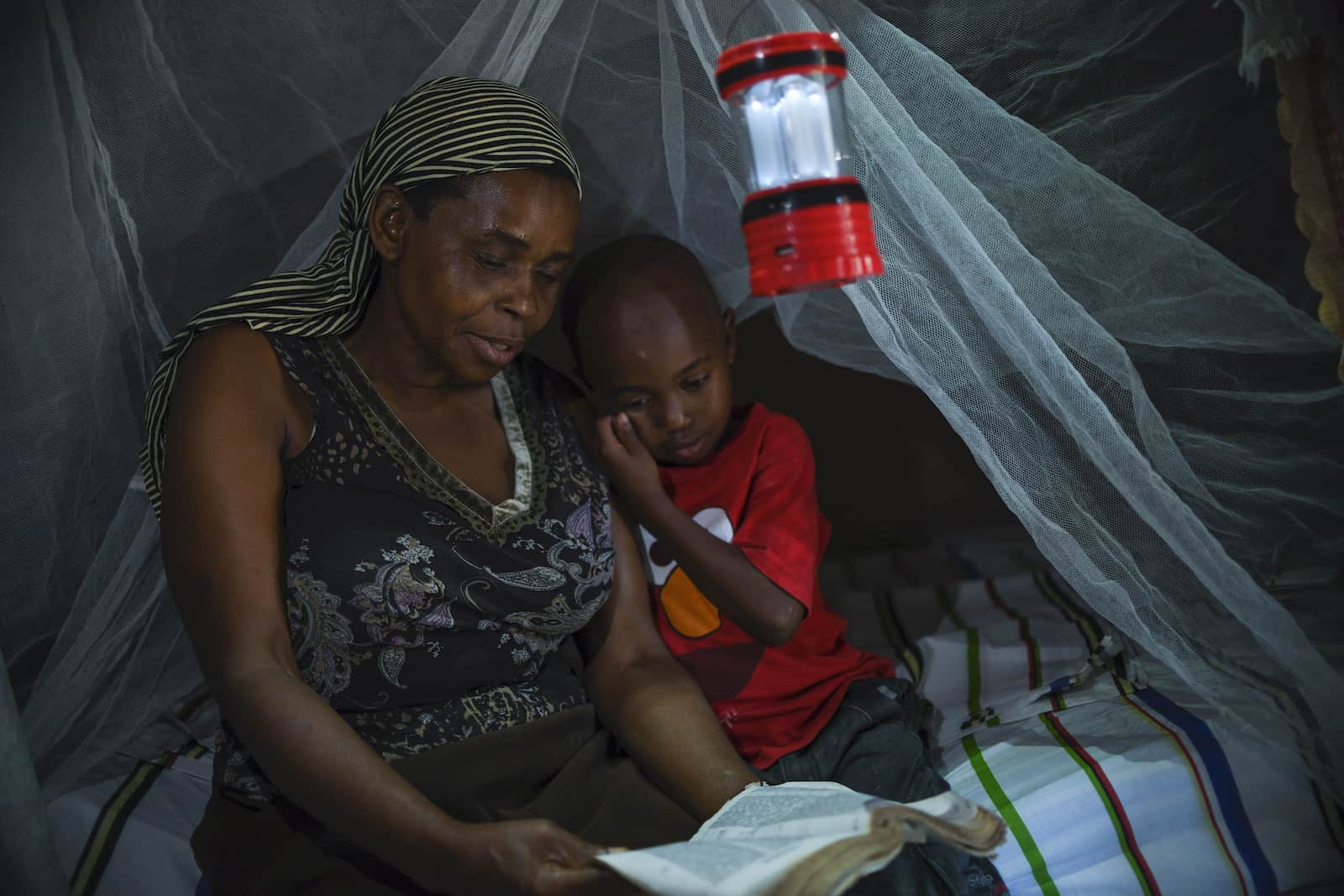 A woman and a boy sit on a bed with a mosquito net and lantern hanging over it, reading the Bible.