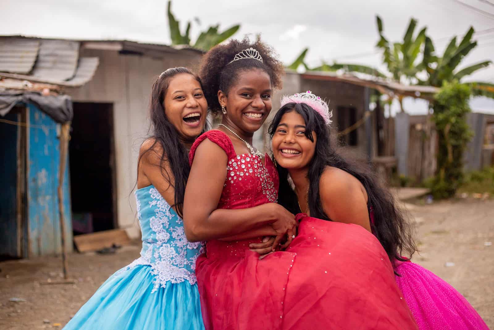Three teenage girls celebrating a birthday tradition, the Quinceañera, in ballgowns hug, smiling and laughing. 