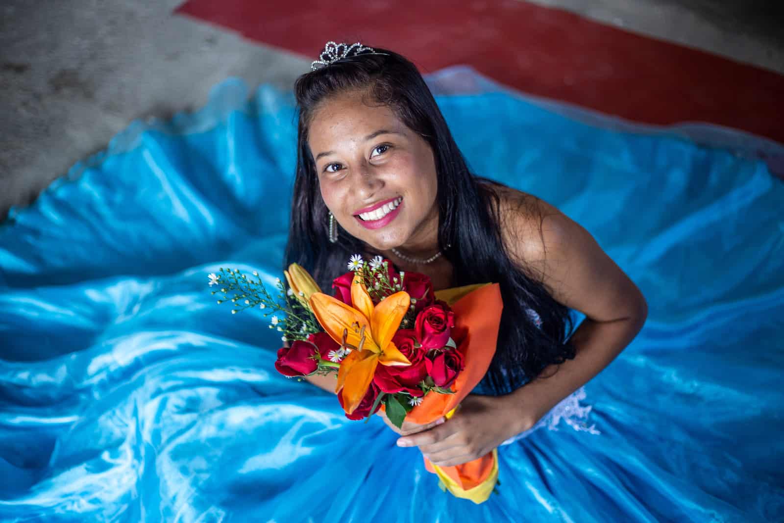 A girl in a blue ballgown sits on the ground, holding a bouquet of flowers.
