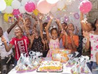 A group of children through confetti in the air, standing in front of a table with a birthday cake on it.