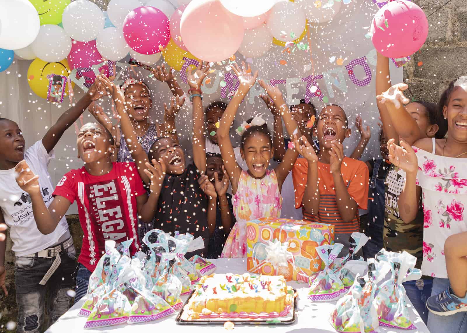 A group of children throw confetti in the air, standing in front of a table with a birthday cake on it. 