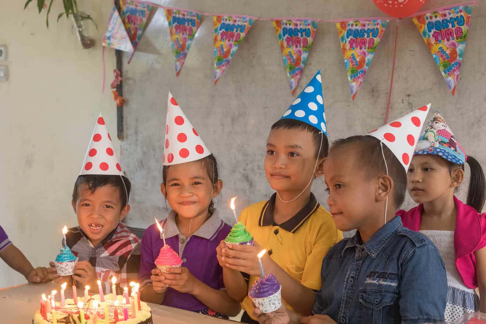 A group of children hold cupcakes, wearing birthday hats and standing in front of a lit birthday cake.