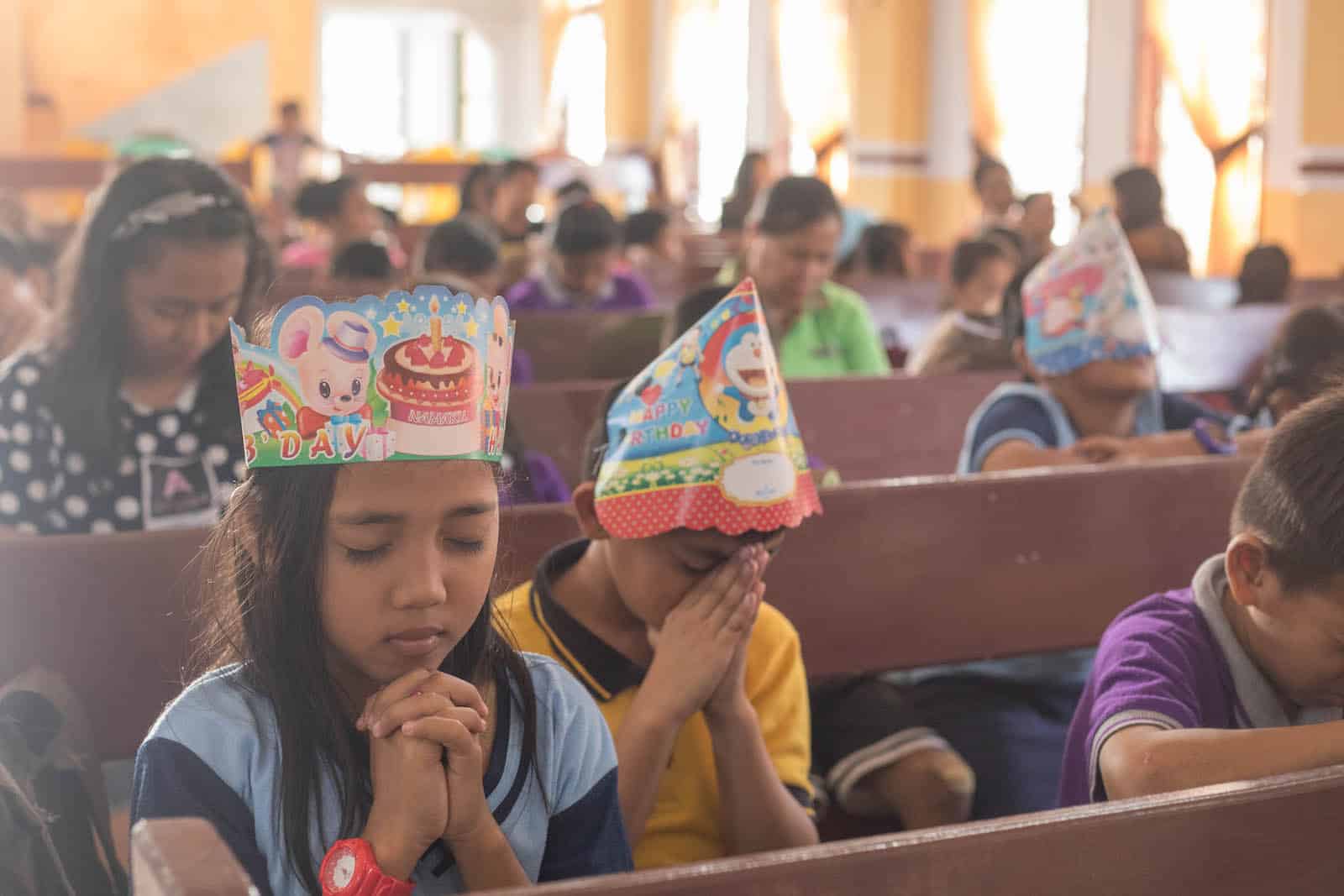 Two children wearing birthday party hats pray, sitting in pews in a church.