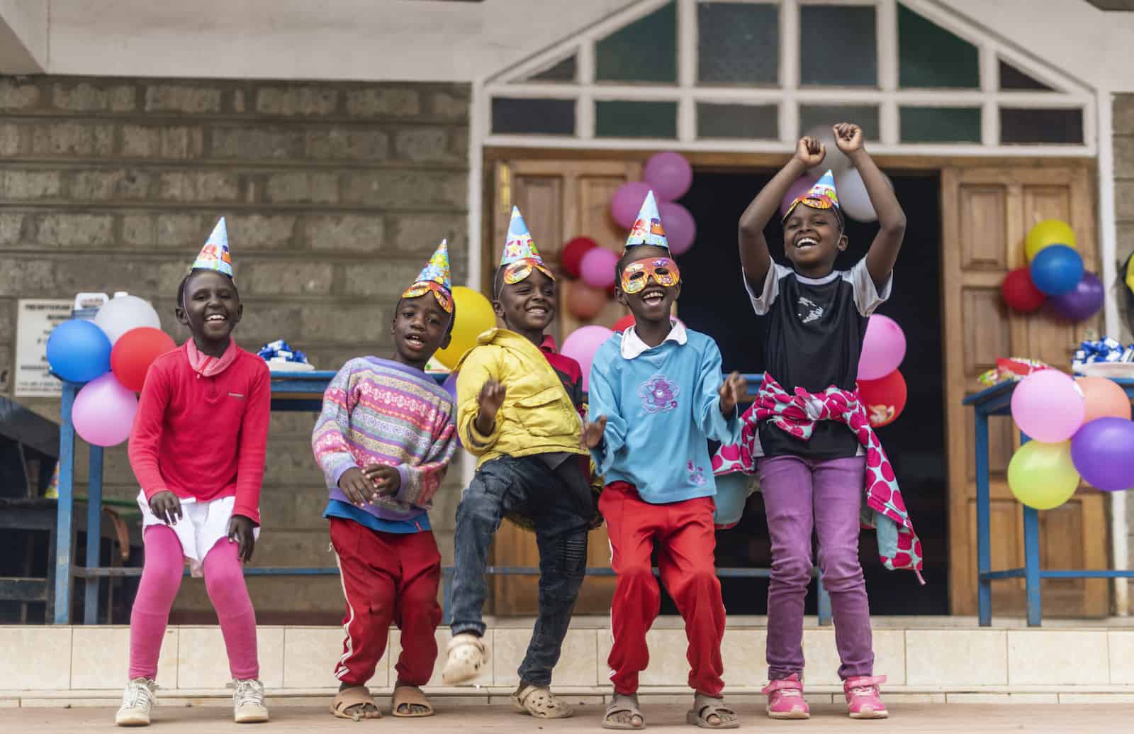 A group of children wearing party hats and party masks smile and dance outside a church.