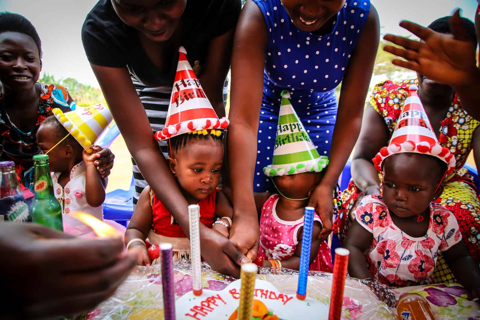 Three babies wearing birthday hats sit in front of a birthday cake. 